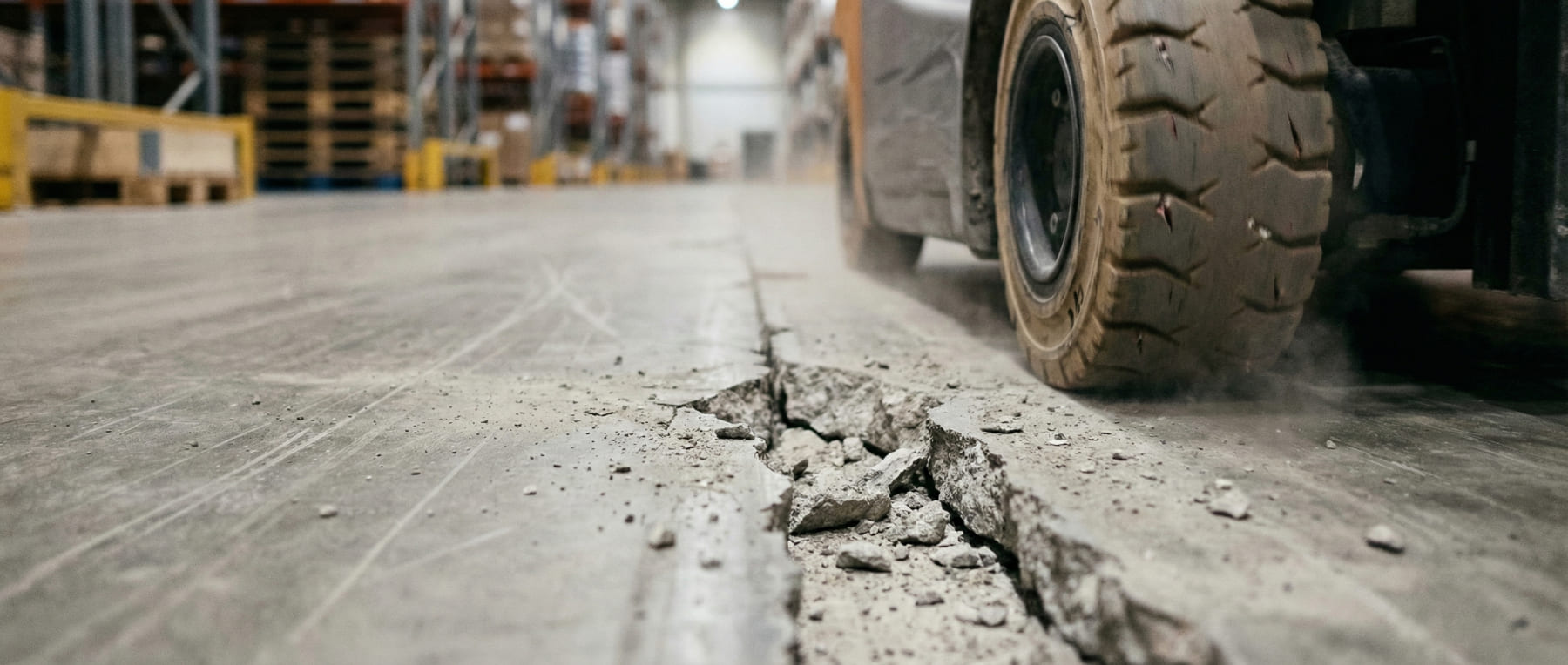 Nahaufnahme eines Gabelstapler-Reifens beim Überfahren einer stark beschädigten Dehnungsfuge mit Kantenabbrüchen - Visualisierung von Verschleiß an Flurförderzeugen durch defekte Industrieböden.