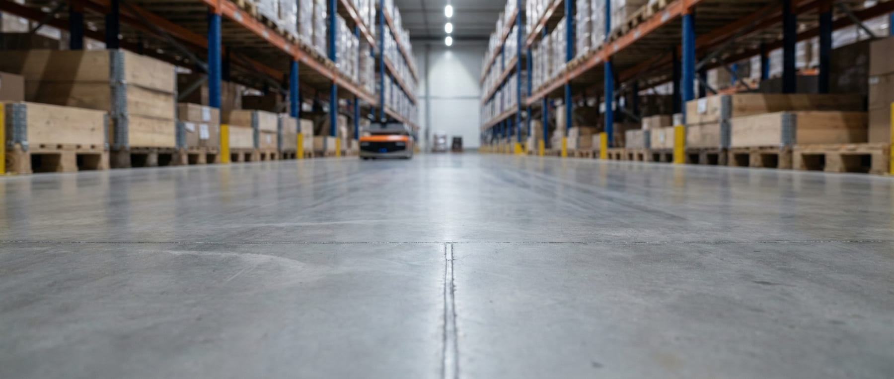 Froschperspektive eines glatten Betonbodens in einem Hochregallager mit sanierten Fugen und einem autonomen Flurförderzeug im Hintergrund - Visualisierung für Logistik und Distribution.