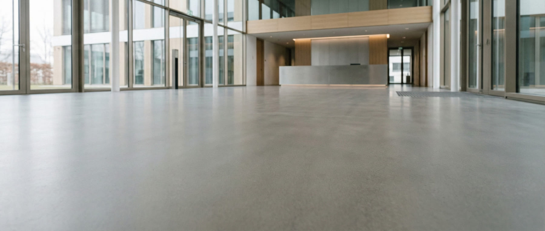 Froschperspektive eines modernen Foyers in einem Bürogebäude mit bodentiefen Fenstern und Empfangstresen - der fugenlose Grindingfloor Sichtestrich unterstreicht die repräsentative Architektur für Office und Verwaltung.